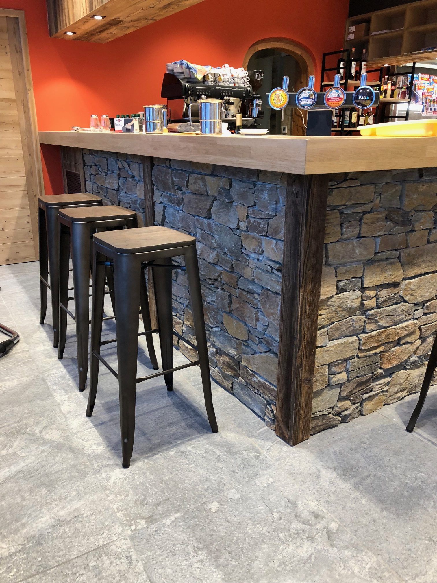 Bar avec façade en pierre, comptoir en bois, tabourets en métal et boissons exposées.