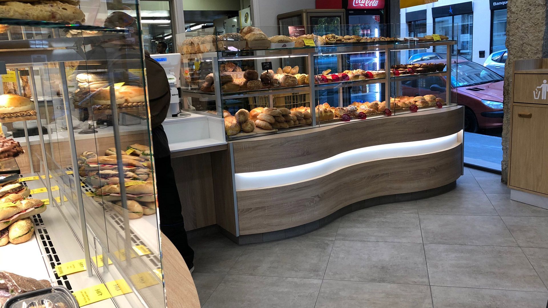 Intérieur d'une boulangerie avec vitrine incurvée présentant des pâtisseries ; vue sur la rue visible à travers la porte.