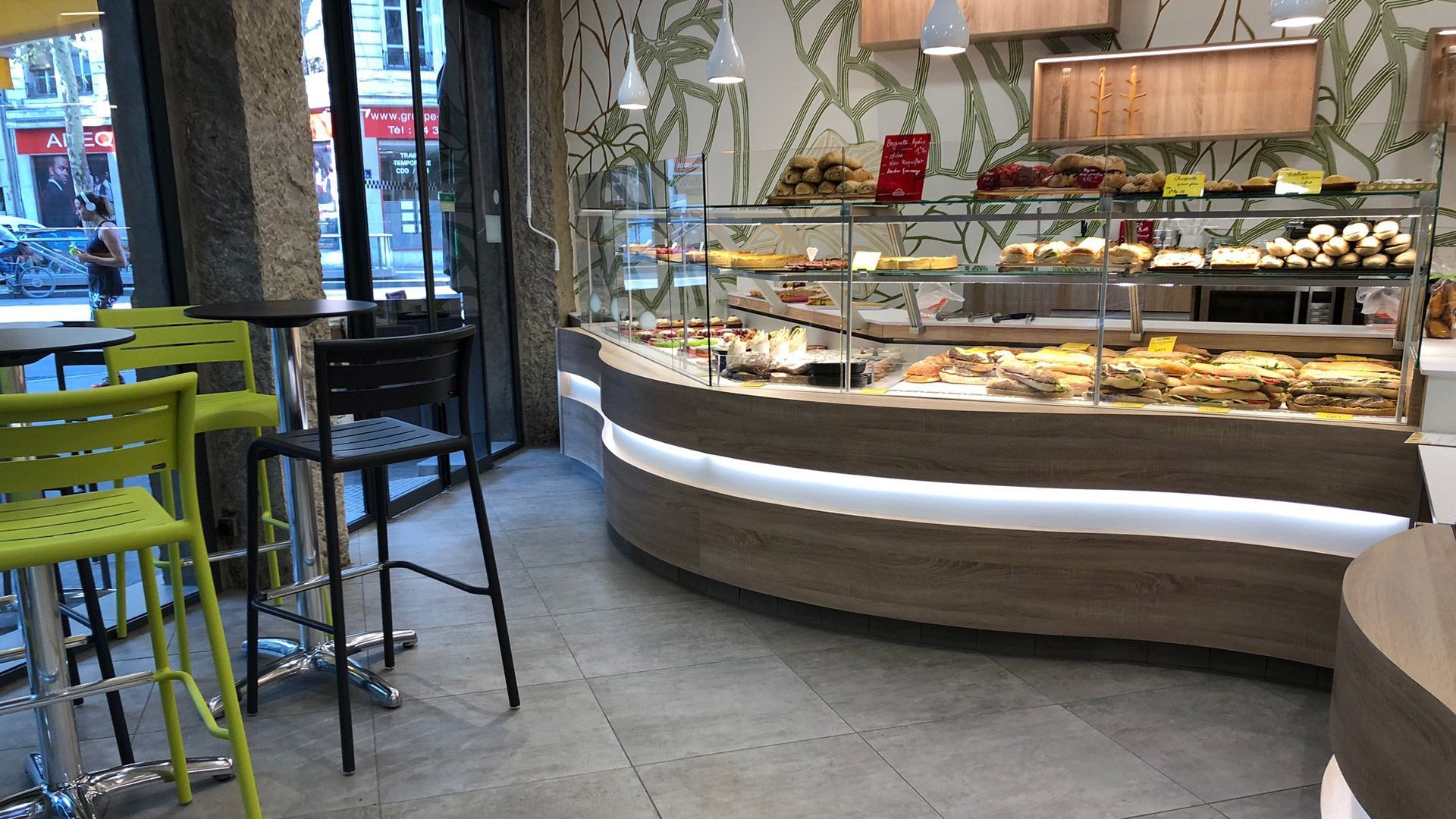 Intérieur d'une boulangerie avec une vitrine de pâtisseries, des tabourets de bar et une fenêtre.