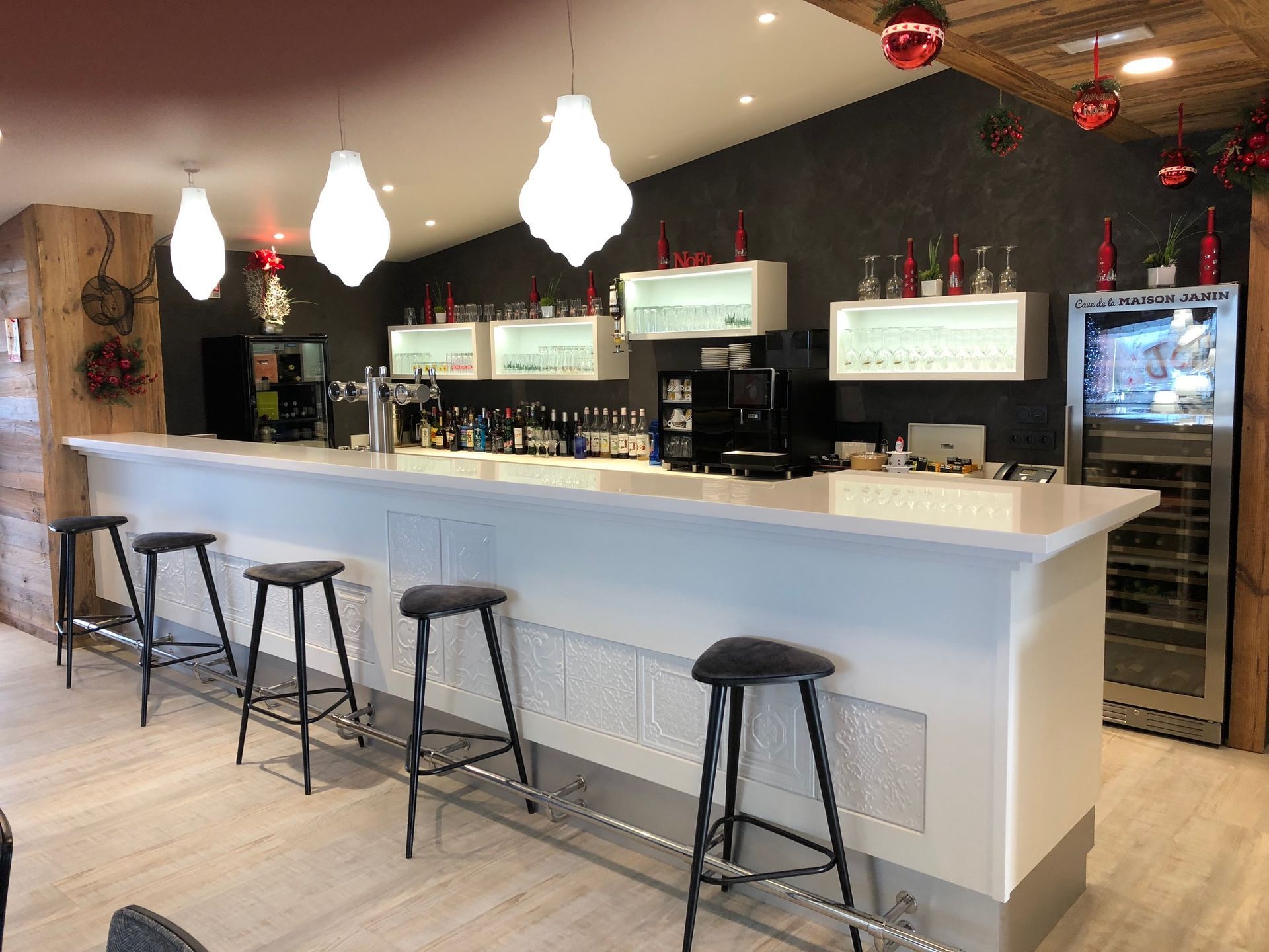 Bar avec comptoir blanc, tabourets noirs, bouteilles d'alcool et luminaires suspendus.