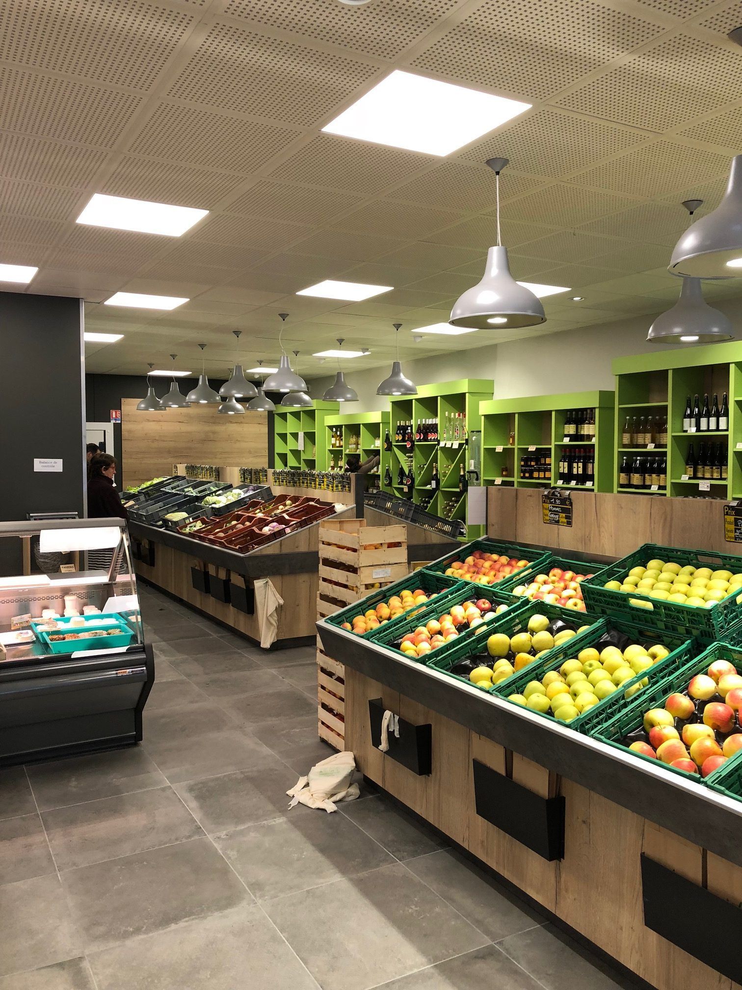 Intérieur d'une épicerie avec des fruits et légumes exposés, sol gris, étagères vertes et suspensions lumineuses.