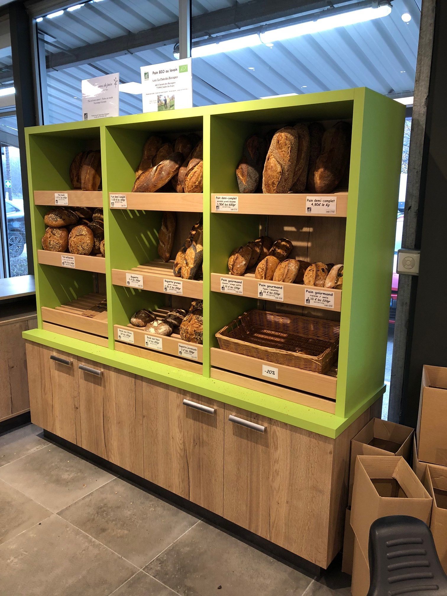 Vitrine de boulangerie présentant différents types de pains à l'intérieur d'une étagère verte avec des placards inférieurs en bois.