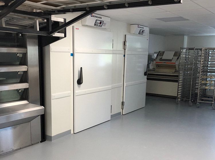 Des réfrigérateurs commerciaux blancs se trouvent dans une boulangerie bien éclairée. D'autres équipements et étagères sont également visibles.