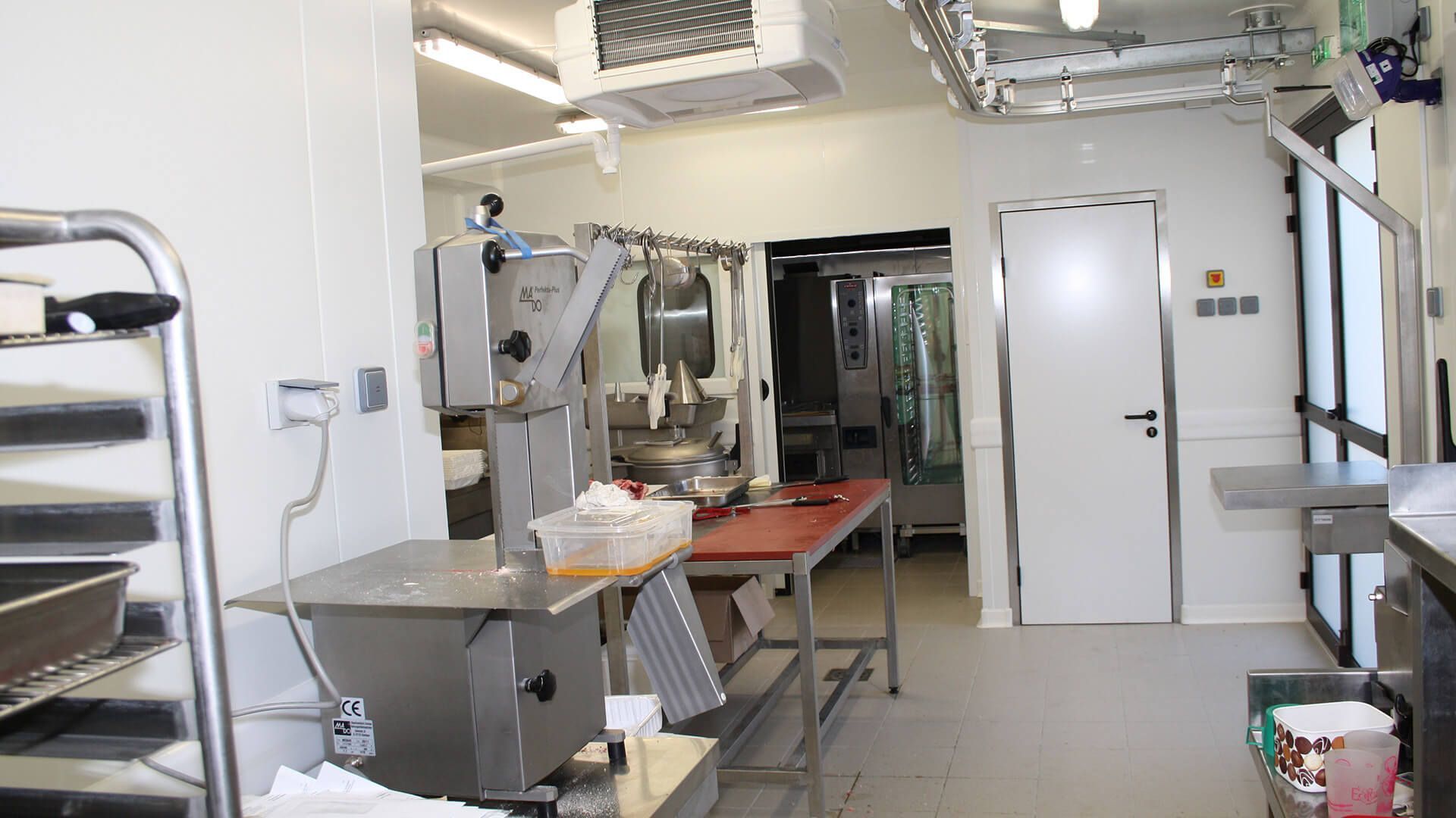 Salle de transformation de la viande : machines en acier inoxydable, table de boucher, viande suspendue et une porte.