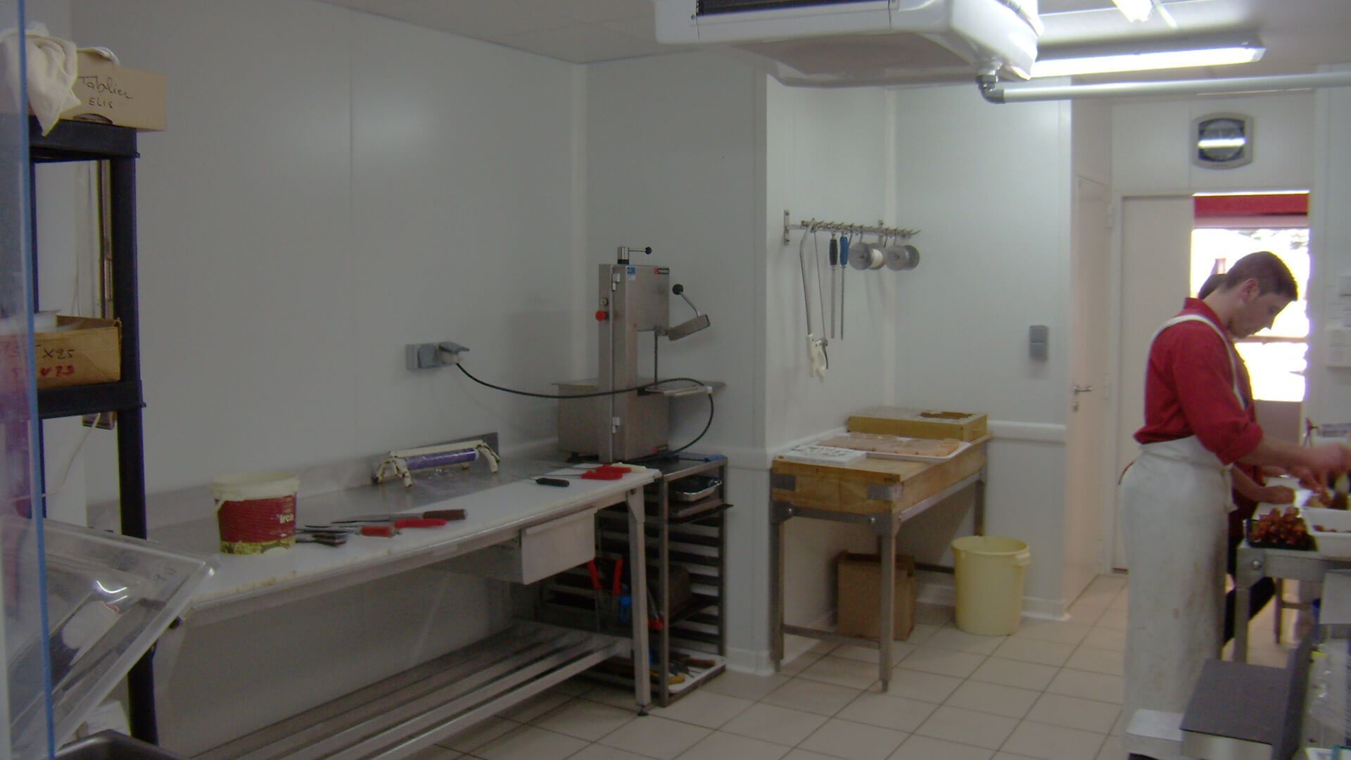 Intérieur de cuisine ; une personne en tablier travaille près de comptoirs en acier inoxydable, à côté d’un hachoir à viande.