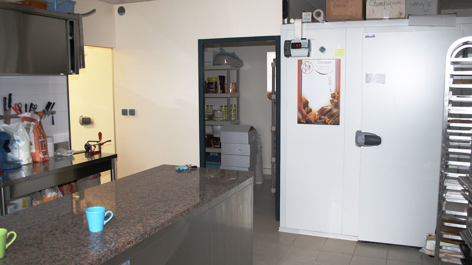 Intérieur de la cuisine avec un plan de travail en inox et un réfrigérateur. Une porte donne accès à un cellier.