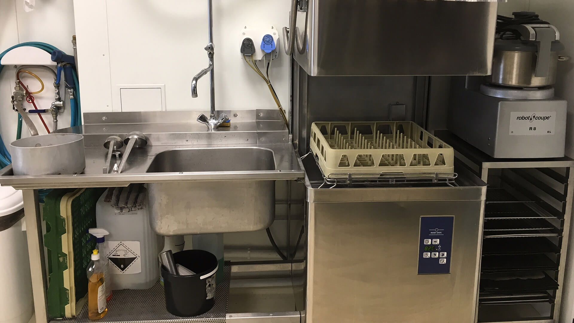 Cuisine professionnelle : évier en acier inoxydable, lave-vaisselle, étagères, produits de nettoyage et robot culinaire.