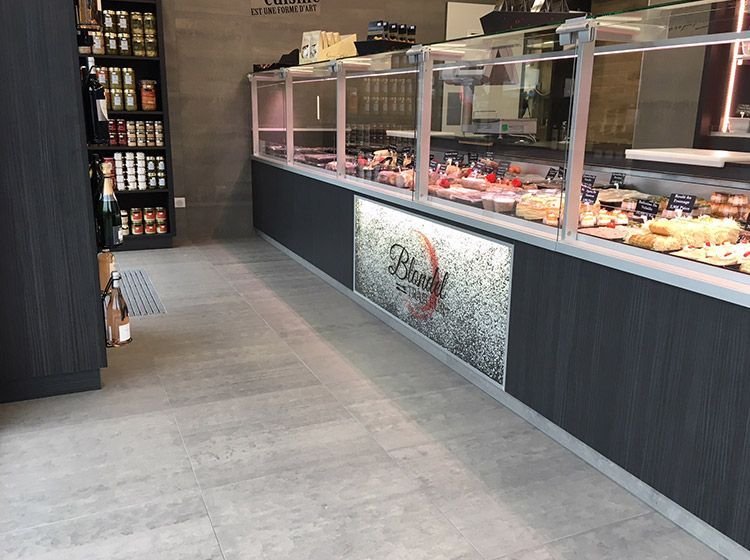 Intérieur d'une épicerie fine, avec une vitrine remplie de produits alimentaires, des étagères garnies et un sol gris.