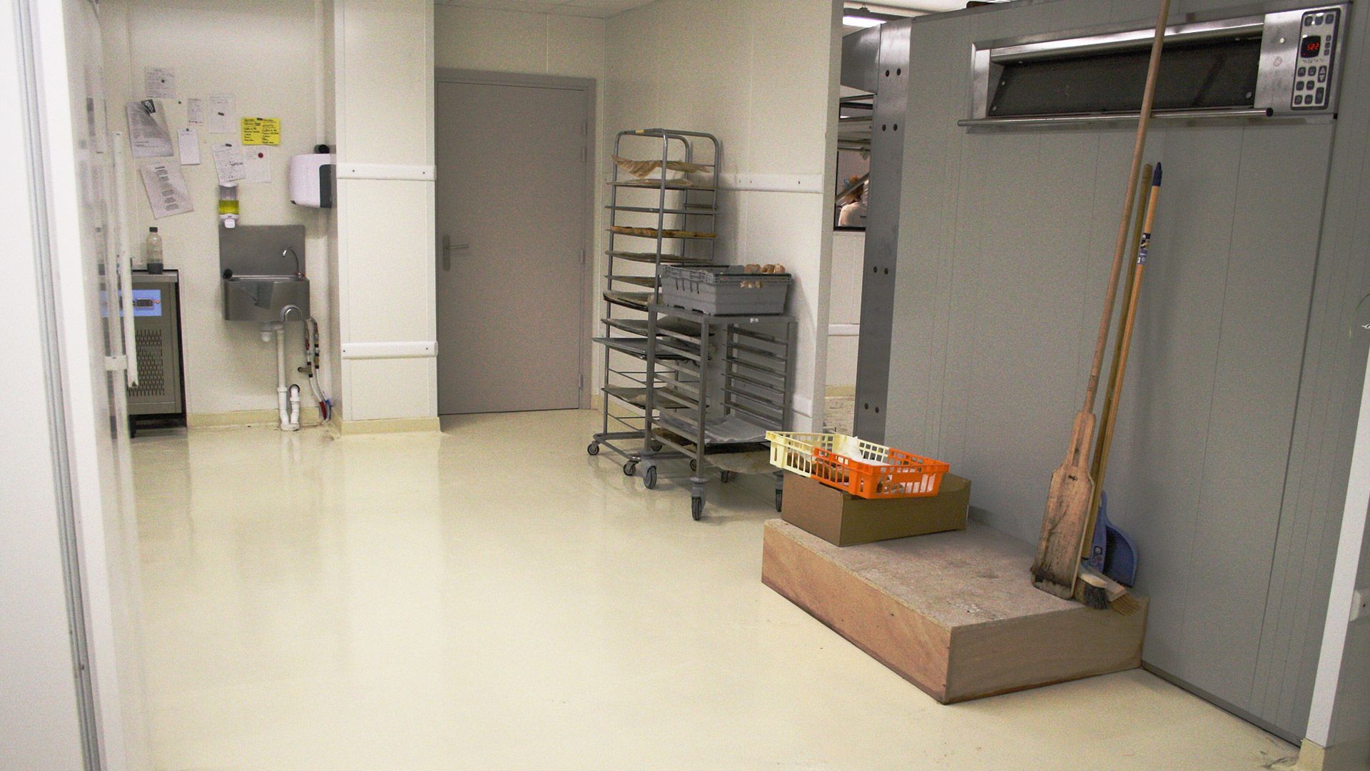 Intérieur d'une cuisine professionnelle. Murs gris, sol clair, porte au centre. Étagères à roulettes, boîtes alimentaires. Balai et serpillière appuyés contre un congélateur.