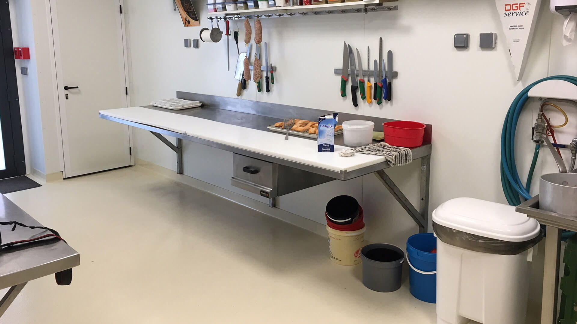 Plan de travail de cuisine en acier inoxydable avec couteaux suspendus, aliments et poubelles.