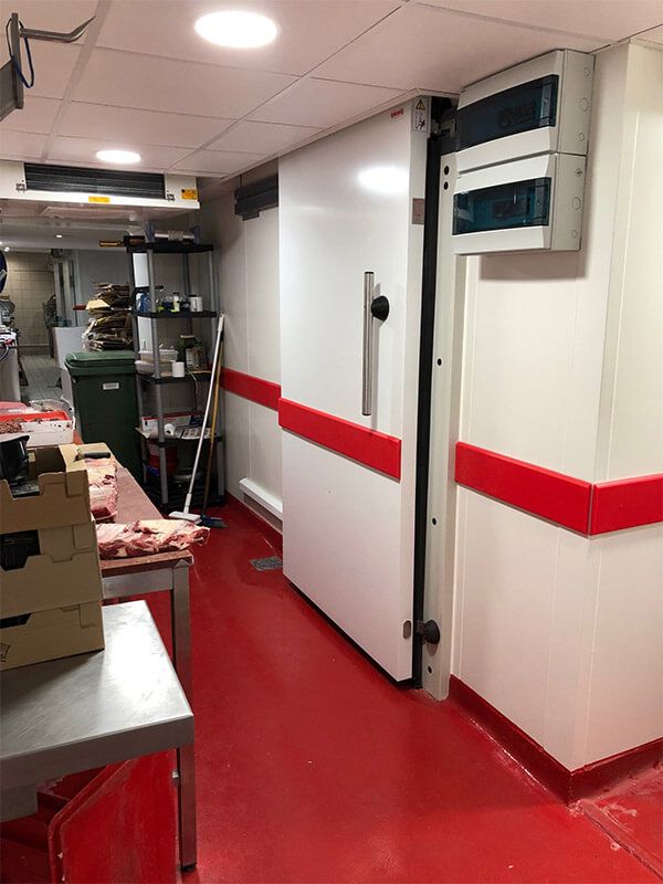 Porte de congélateur commercial blanche avec des touches de rouge dans une cuisine au sol rouge.