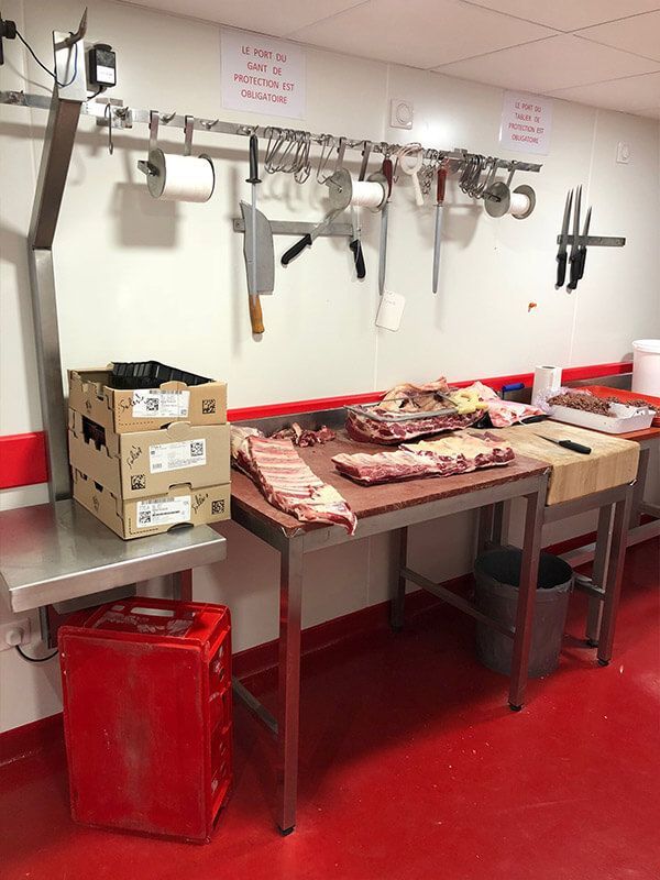 Espace de travail d'une boucherie : viande sur une table en inox, couteaux et boîtes sur le comptoir. Sol rouge.