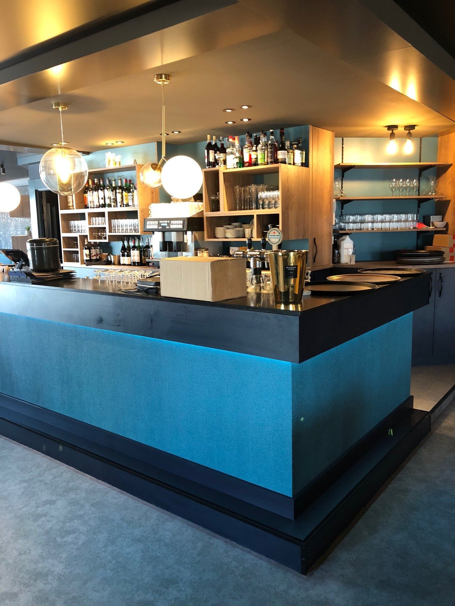 Un bar à façade turquoise avec des bouteilles d'alcool, de la verrerie et des suspensions lumineuses en forme de globe, probablement dans un restaurant.