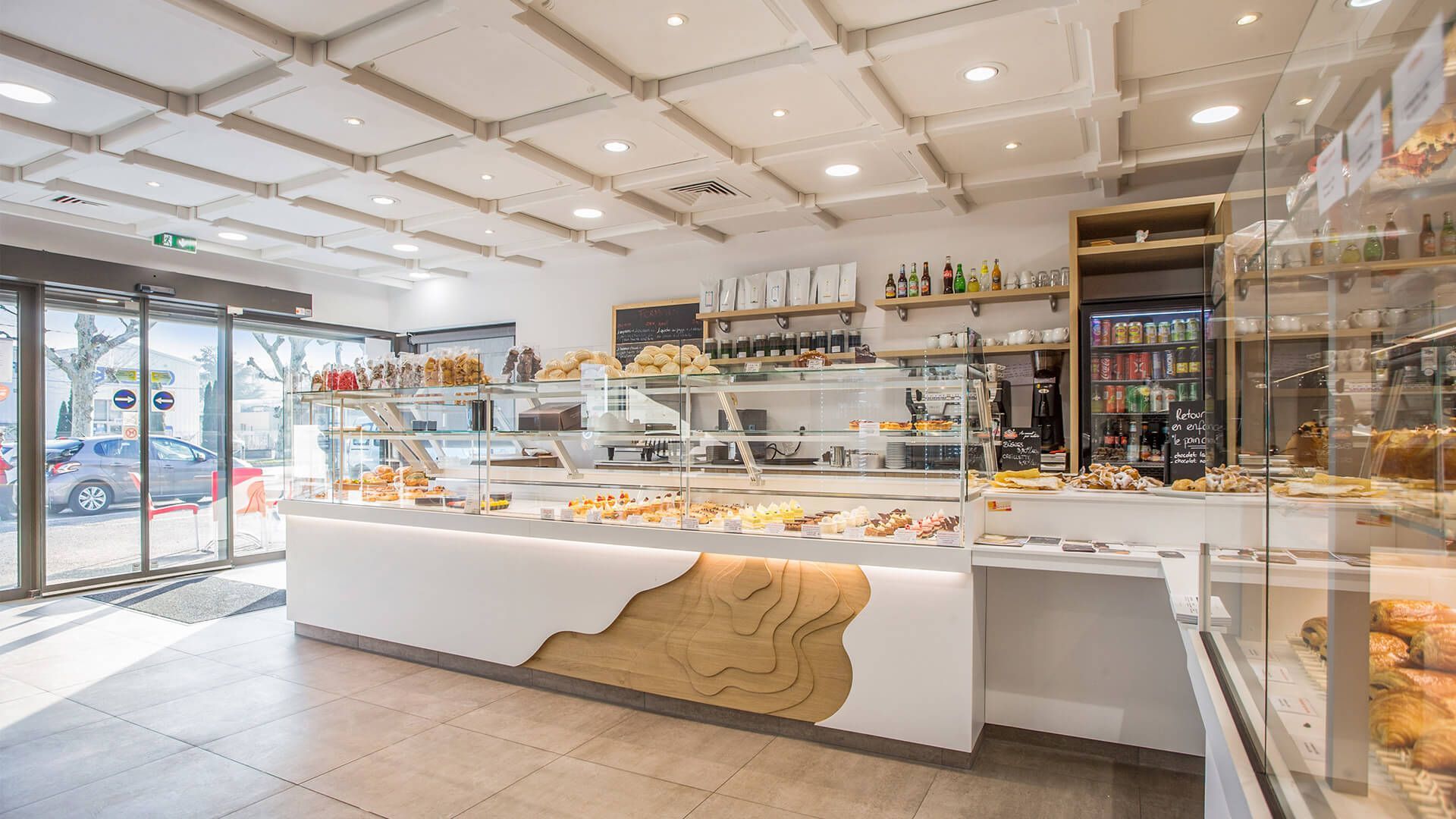 Intérieur de la boulangerie aux tons blancs et boisés ; vitrines en verre présentant les pâtisseries.