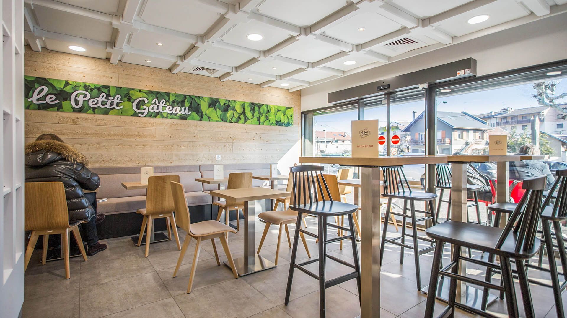 Intérieur du café Le Petit Gillou avec tables, chaises et fenêtre. Une personne est assise dans une banquette.