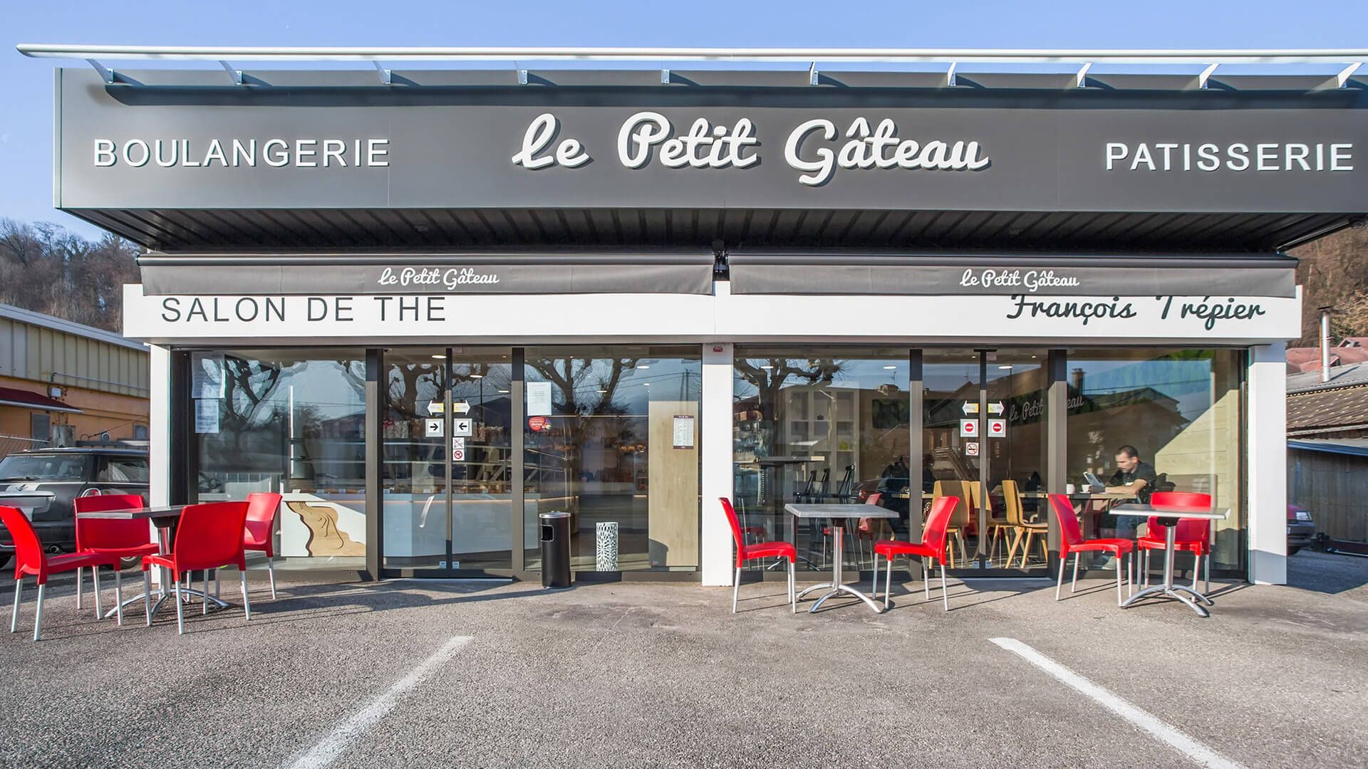Boulangerie « Le Petit Gâteau » avec auvent. Chaises et tables rouges sur la terrasse.