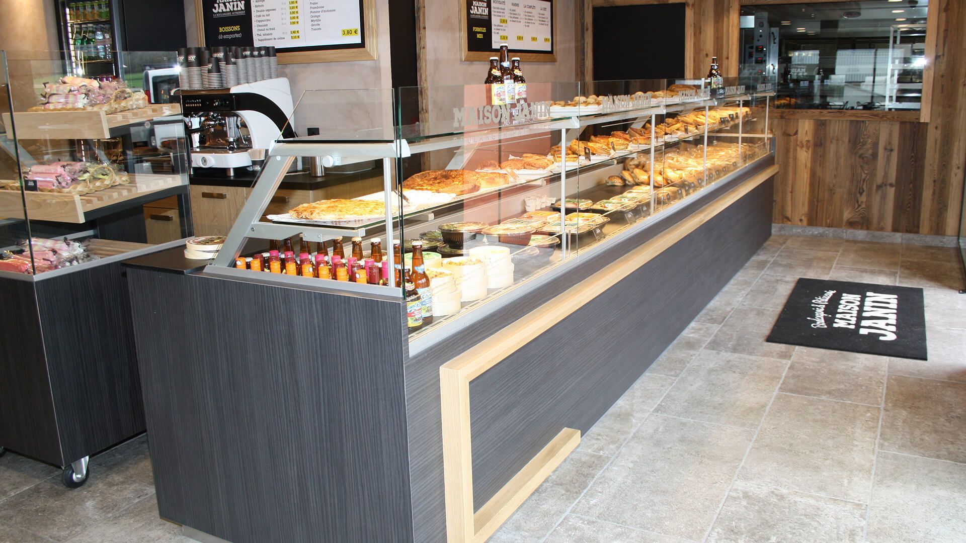 Comptoir de boulangerie avec pâtisseries. Tons gris et beige, sol carrelé.