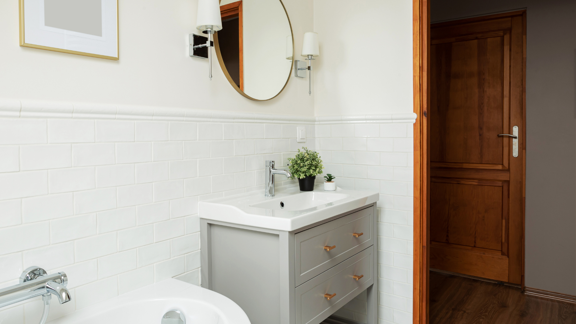 Salle de bain avec carrelage blanc, vanité grise, miroir et appliques dorés et porte en bois.