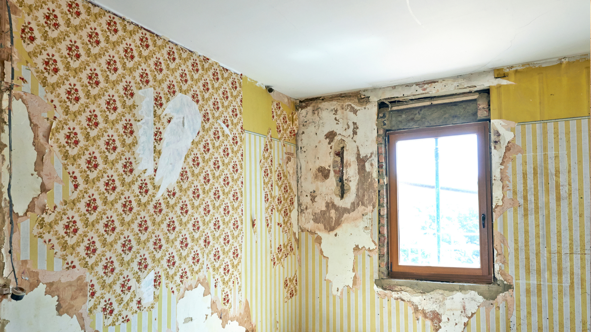 Chambre avec papier peint à motifs floraux et à rayures qui s'écaille autour d'une fenêtre.