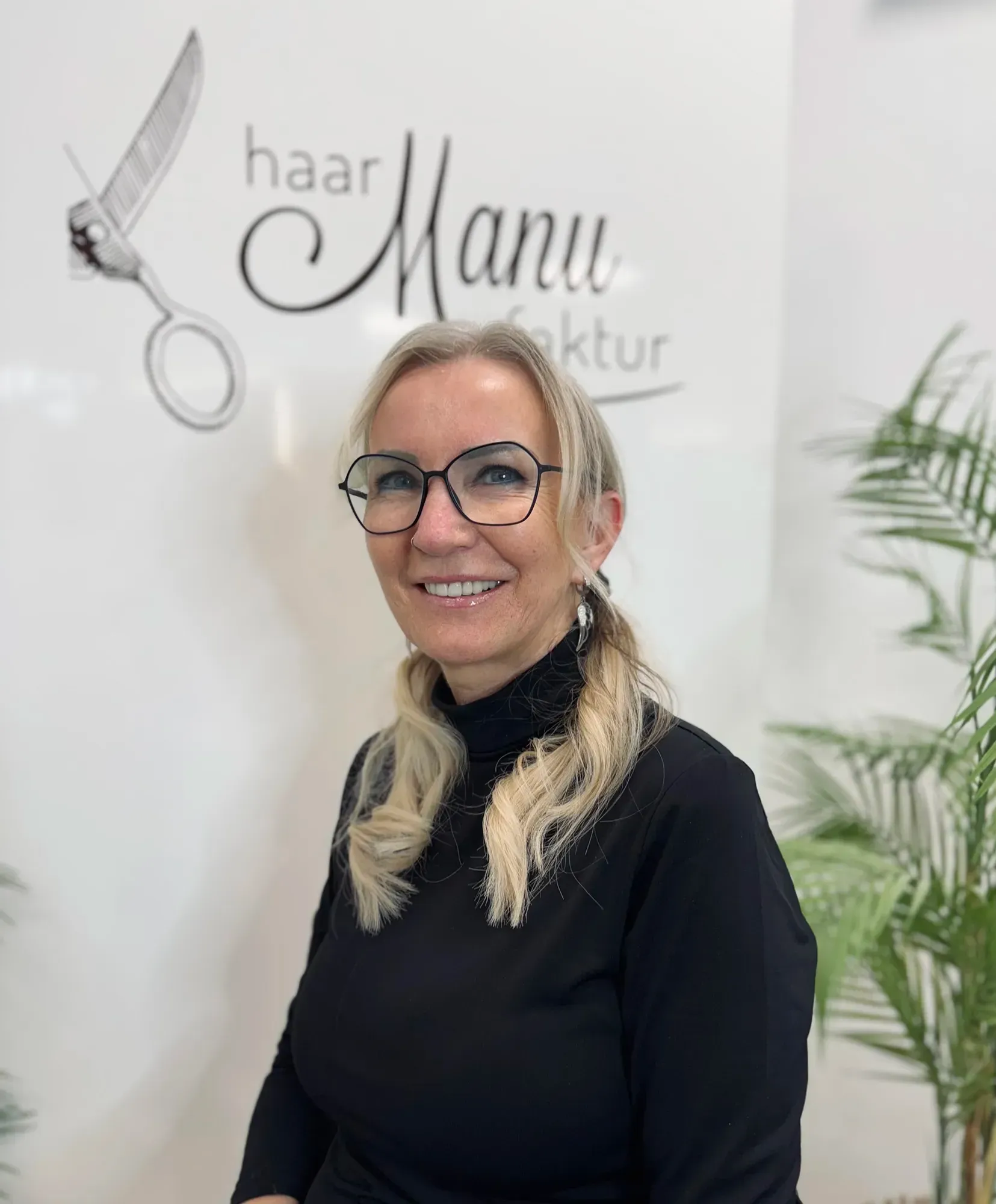 Eine Frau mit Brille lächelt vor einem Schild mit der Aufschrift „Haar Manu“
