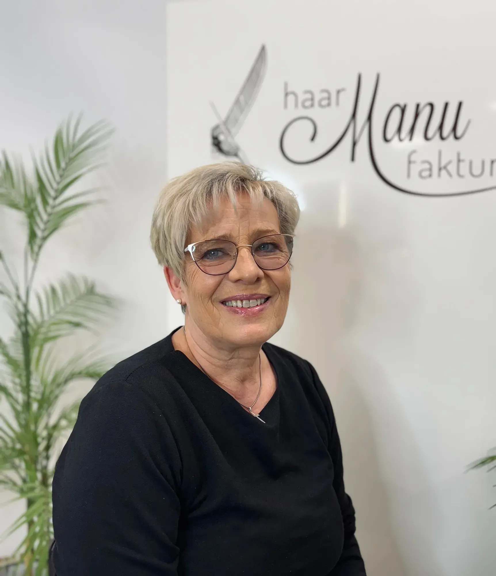 Eine Frau mit Brille lächelt vor einem Schild mit der Aufschrift „Haar Manu Faktur“