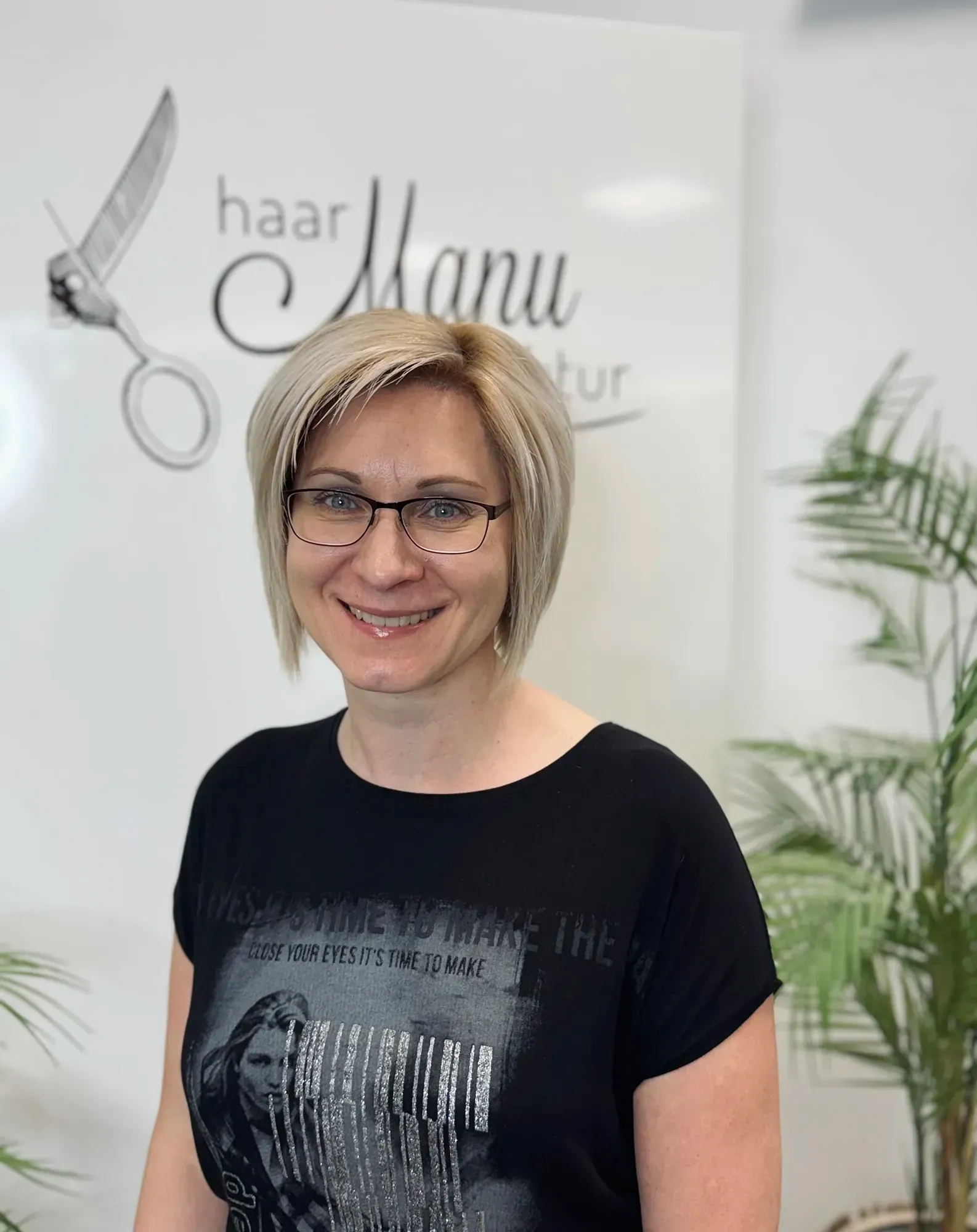 Eine Frau mit Brille und schwarzem Hemd lächelt vor dem Schild eines Friseursalons.