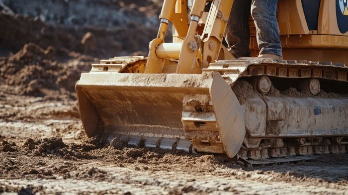 Un bulldozer jaune pousse de la terre sur un chantier. On aperçoit les jambes et les bottes d'une personne.