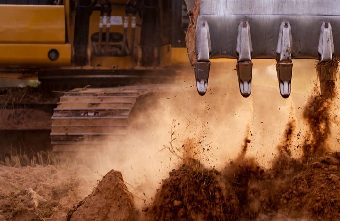 Une excavatrice creuse la terre, soulevant un nuage de poussière. Machine jaune, gros plan sur les dents du godet.