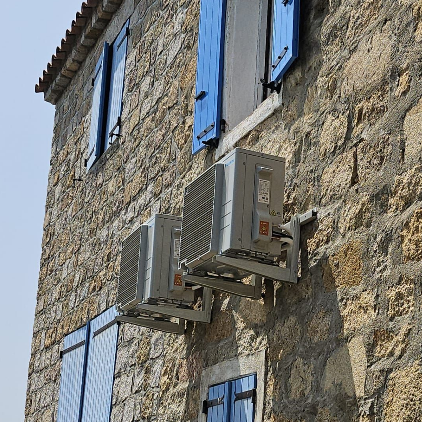 Mur en pierre avec volets bleus et deux unités de climatisation fixées sous une fenêtre.