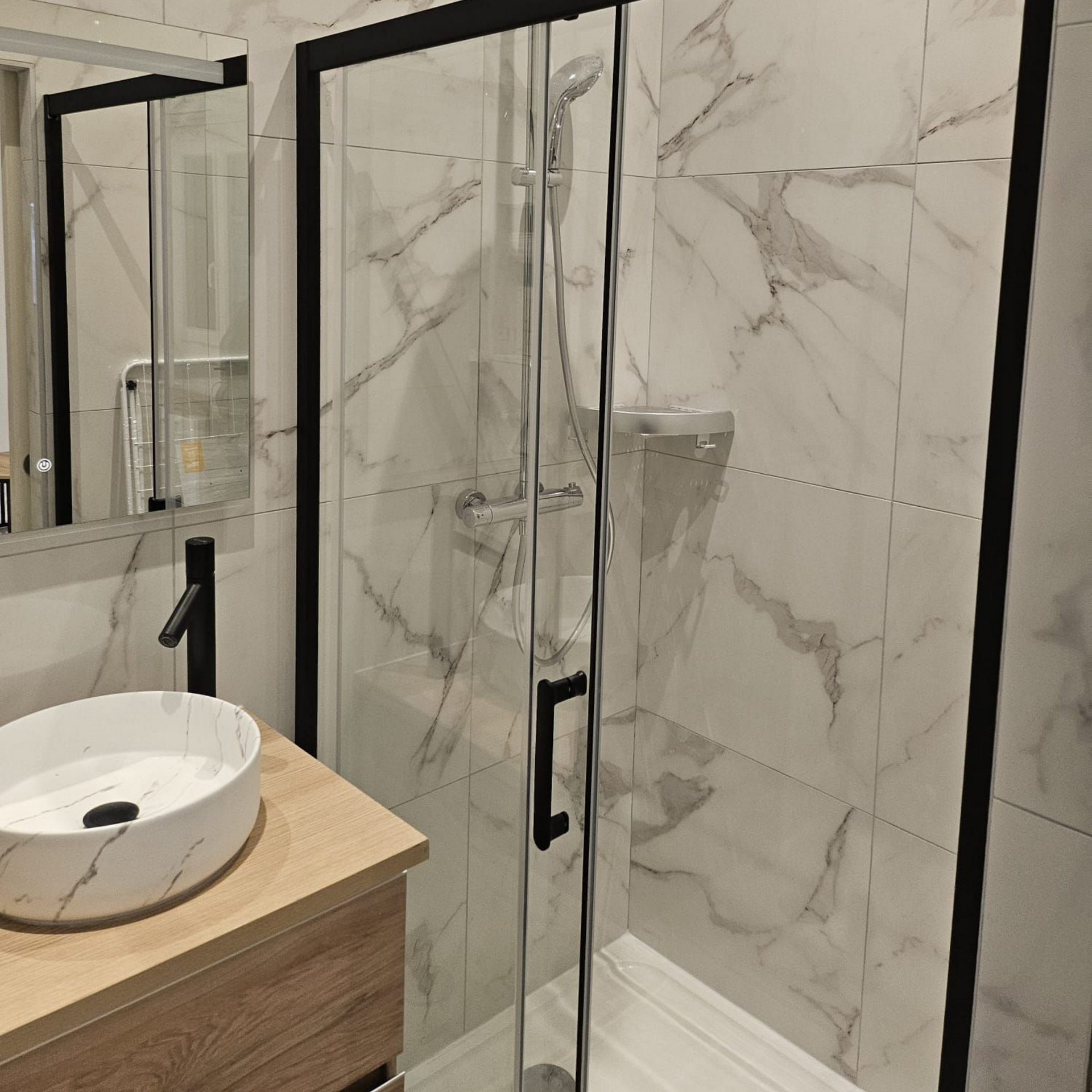 Salle de bains moderne avec douche en marbre, portes vitrées à cadre noir et lavabo rond sur meuble lavabo en bois.