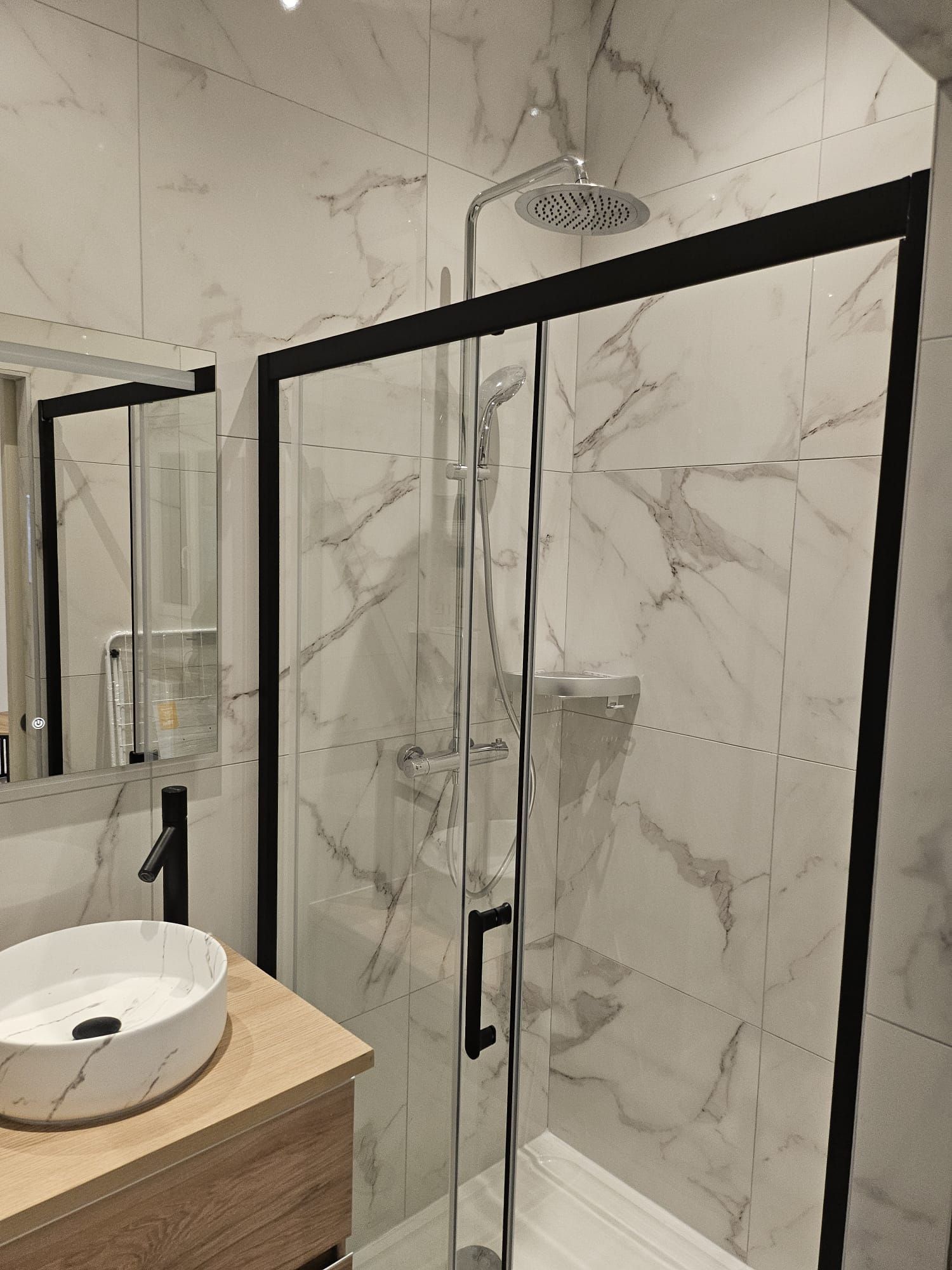 Douche de salle de bains moderne avec portes en verre à cadre noir, murs carrelés en marbre et lavabo au premier plan.