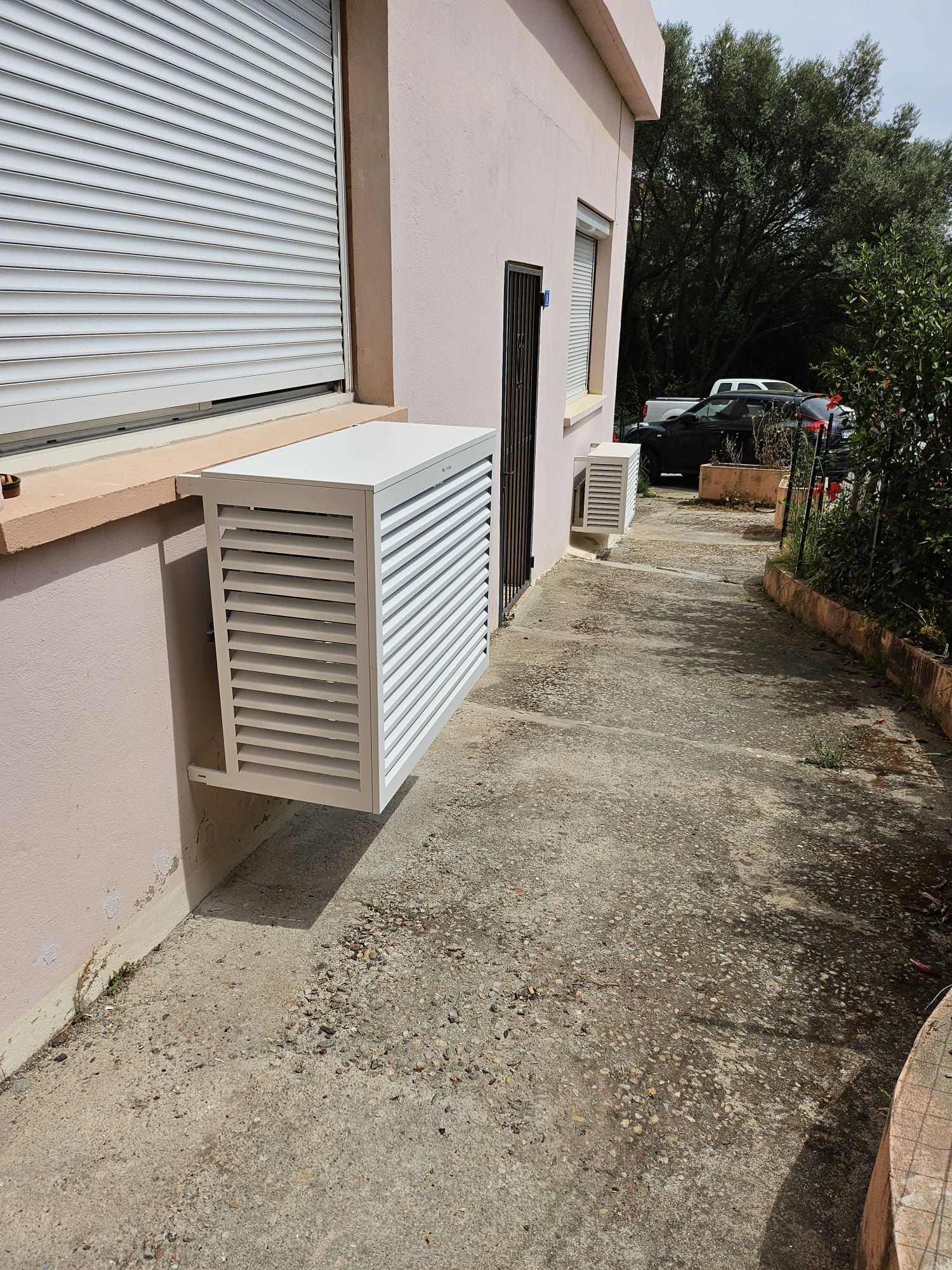 Allée latérale extérieure longeant un bâtiment beige avec un boîtier utilitaire blanc à persiennes et des volets fermés.