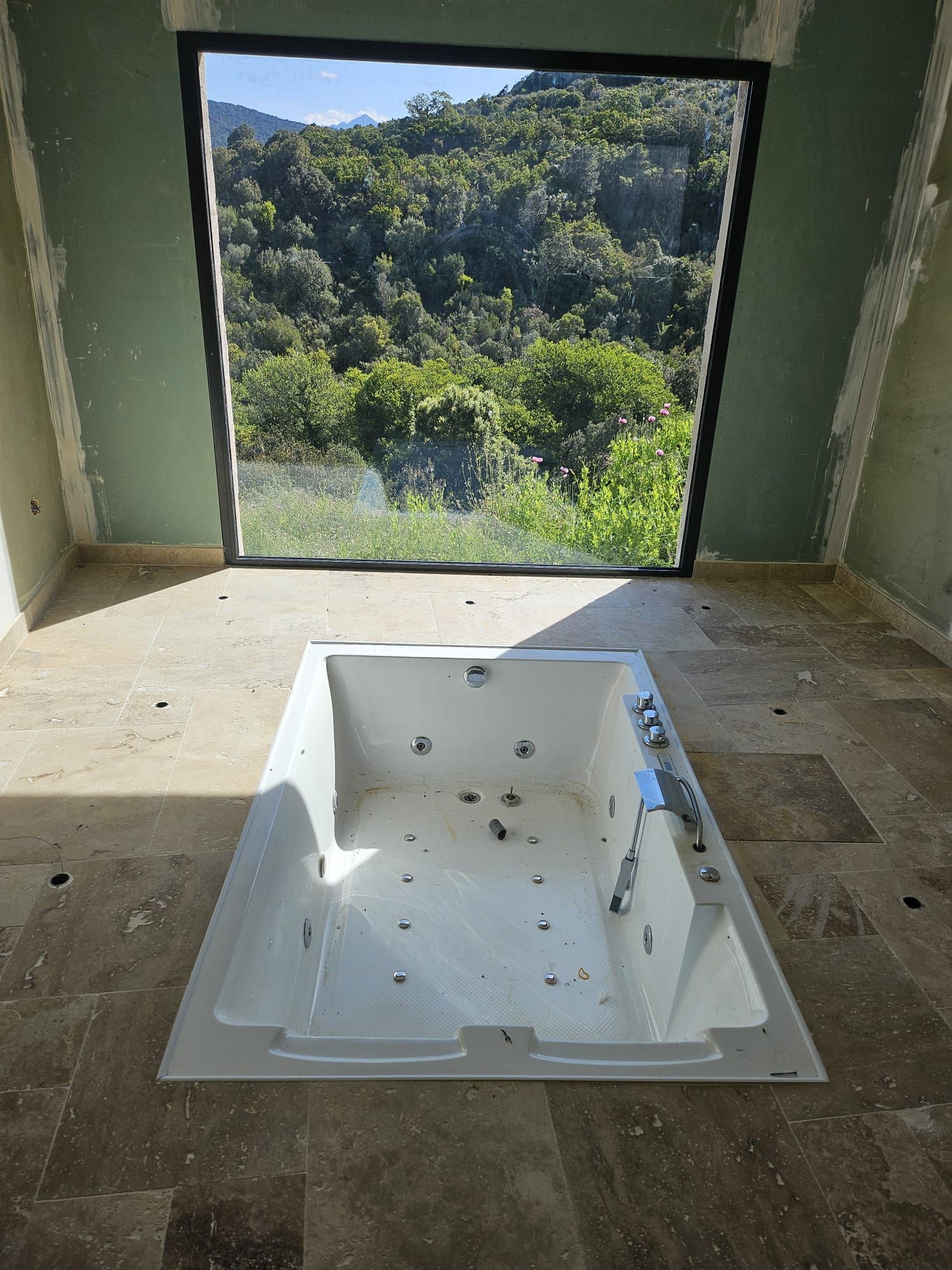 Salle de bains inachevée avec une baignoire blanche placée au centre, devant une grande fenêtre ouverte donnant sur les arbres.
