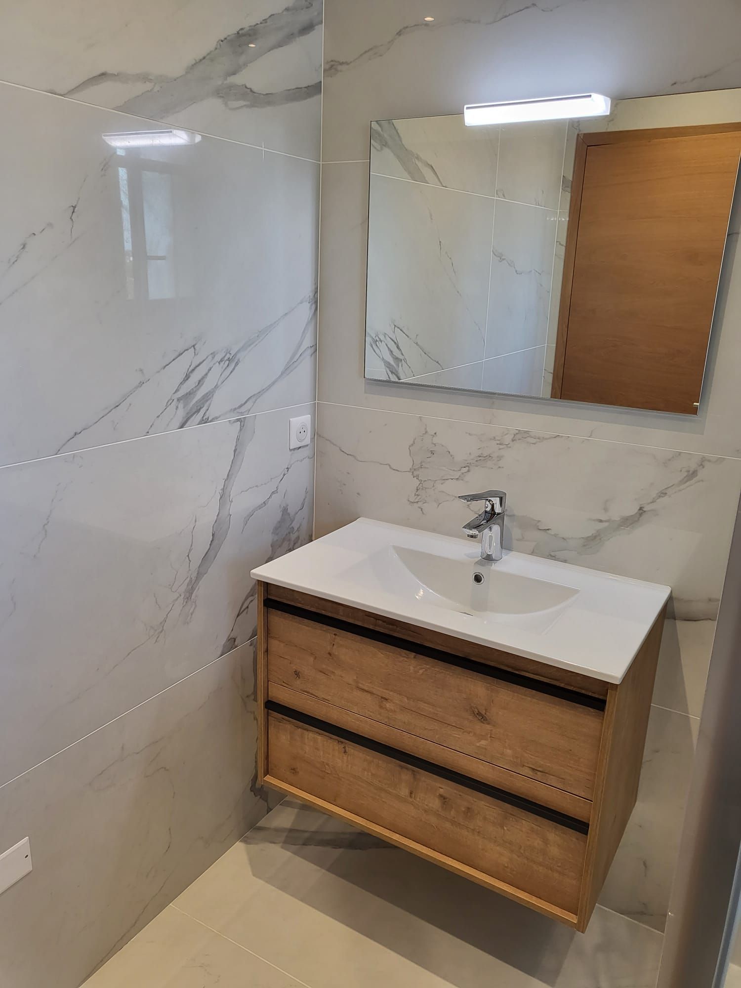 Meuble lavabo moderne avec carrelage imitation marbre, armoire en bois, lavabo blanc et miroir mural.