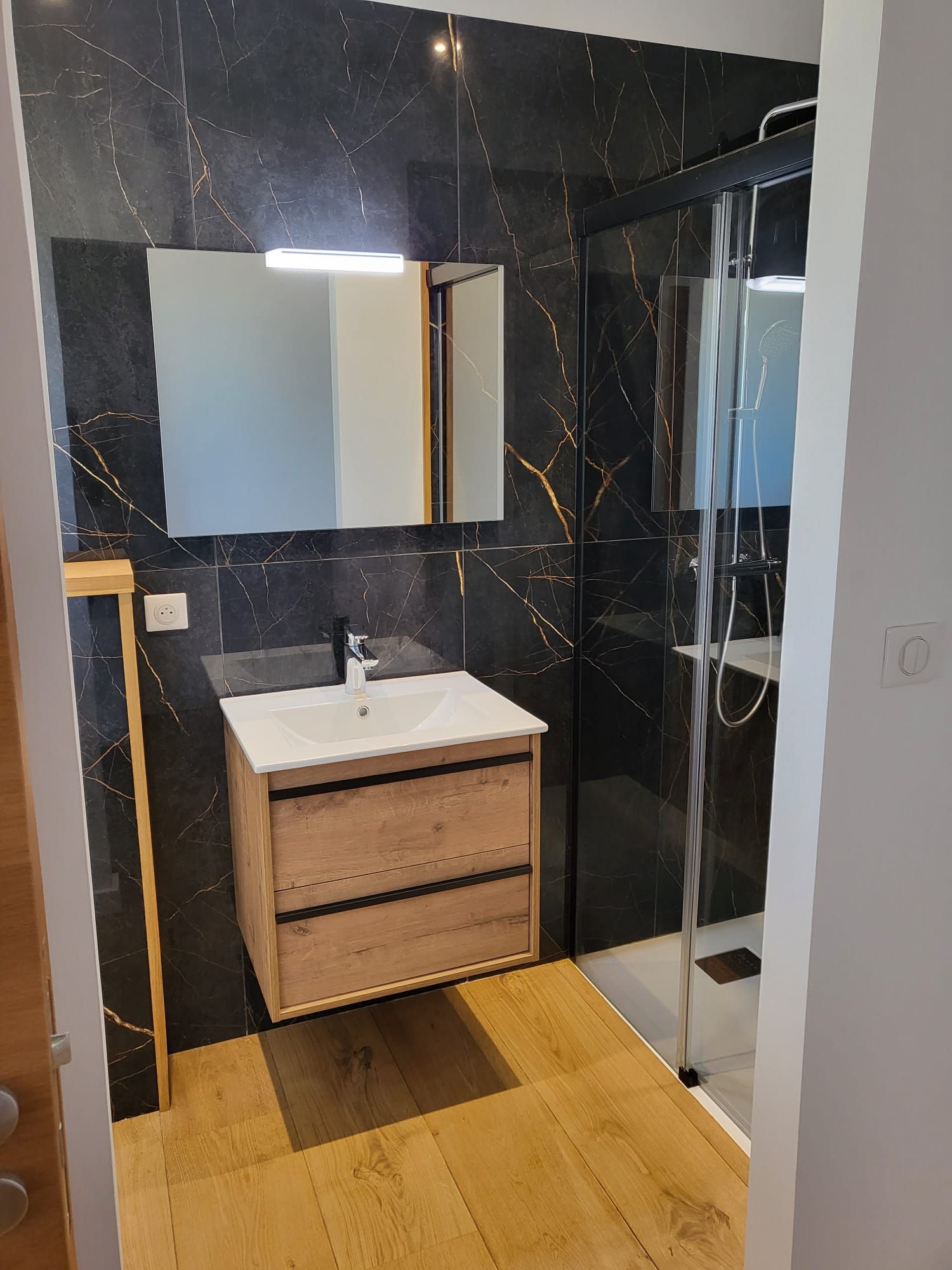 Salle de bains moderne avec murs en marbre noir, meuble lavabo en bois, miroir, lavabo et cabine de douche en verre.