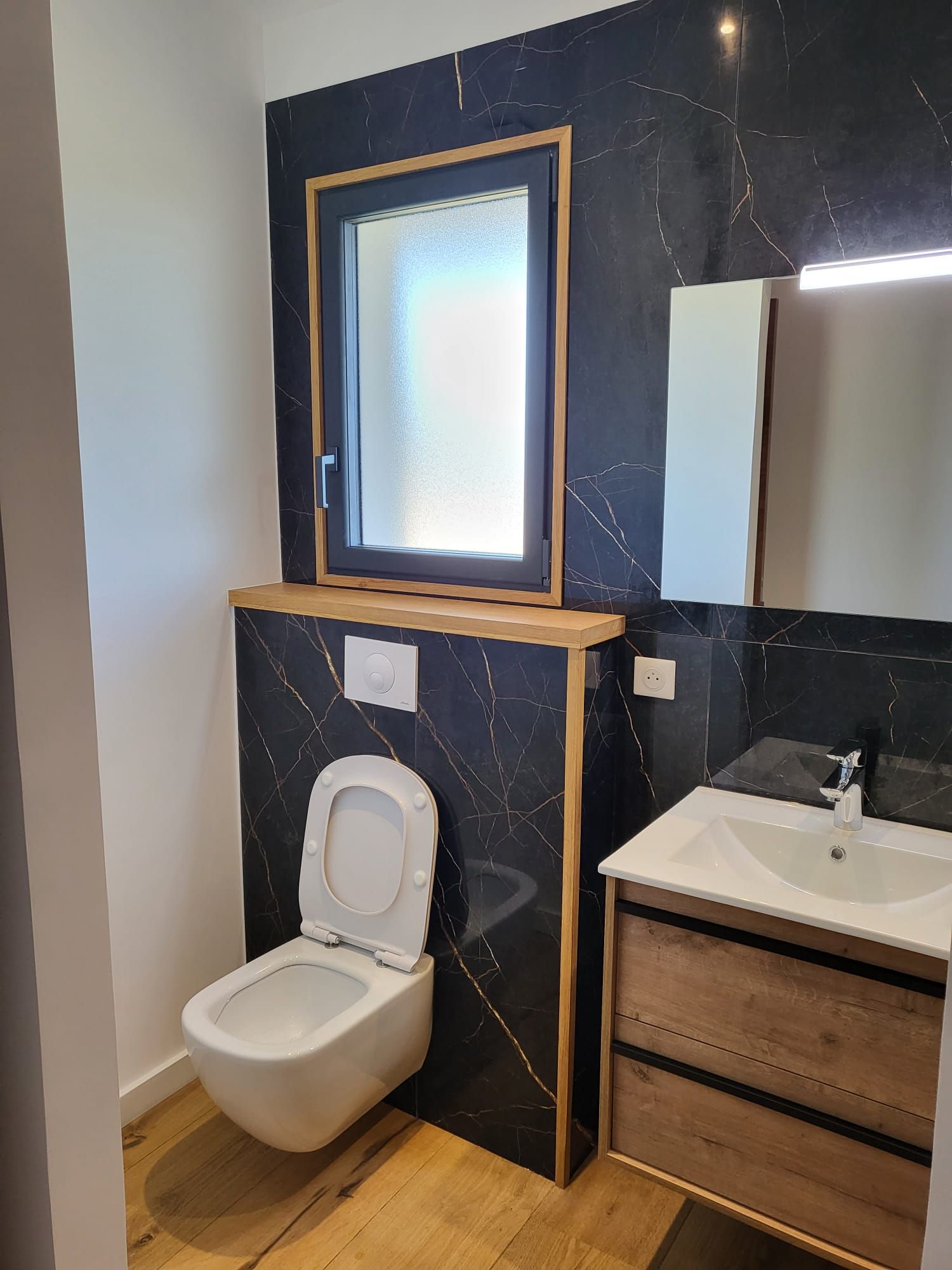 Petite salle de bains avec toilettes suspendues, meuble lavabo, miroir et fenêtre dans un mur carrelé de noir.