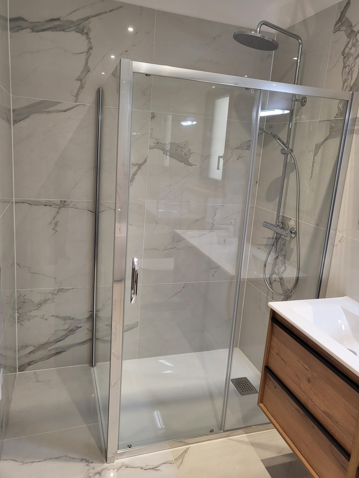 Cabine de douche en verre avec cadre chromé dans une salle de bains carrelée en marbre, à côté d'un meuble lavabo en bois.