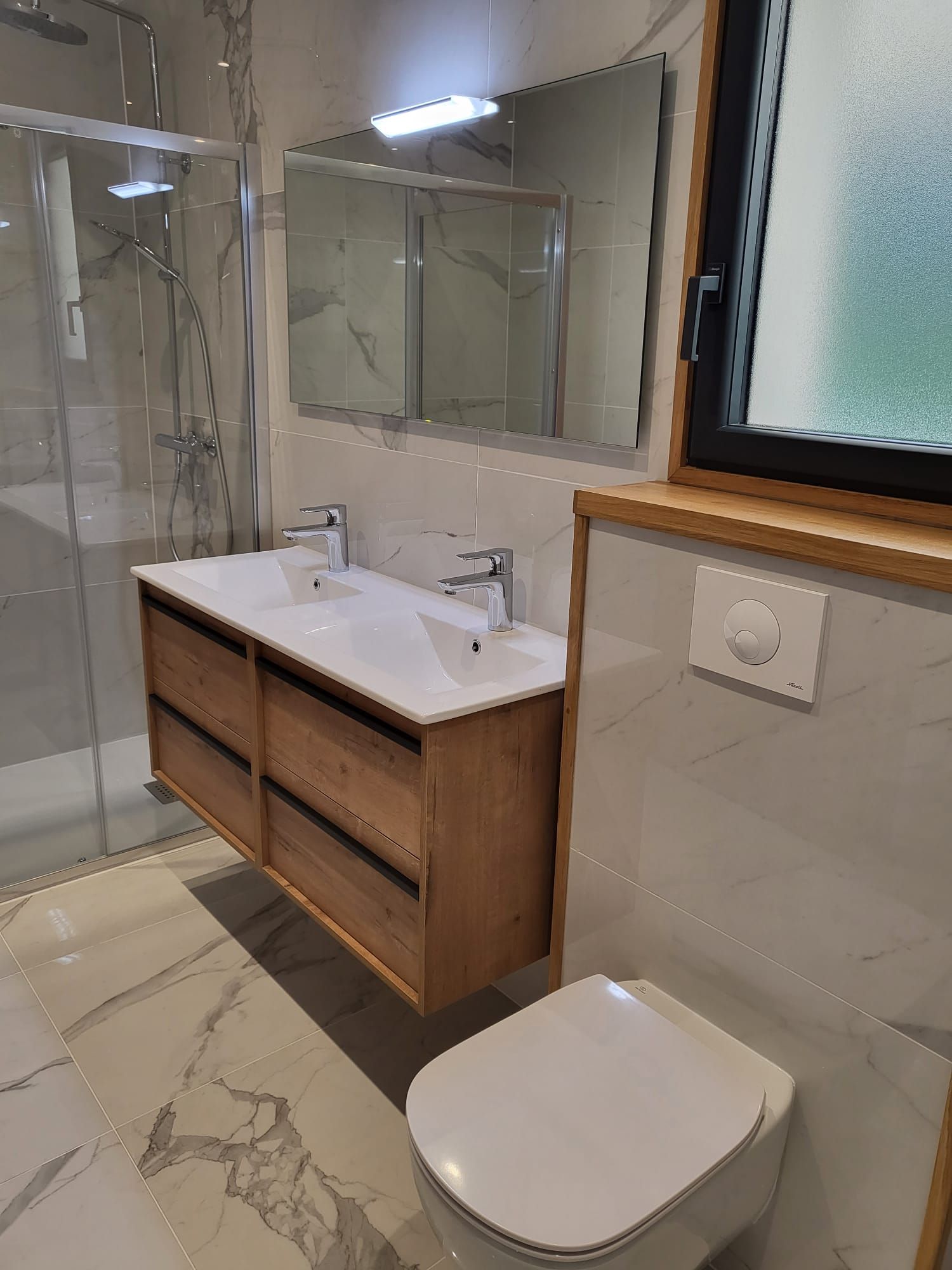 Salle de bains moderne avec douche en verre, meuble lavabo en bois, miroir et toilettes suspendues.
