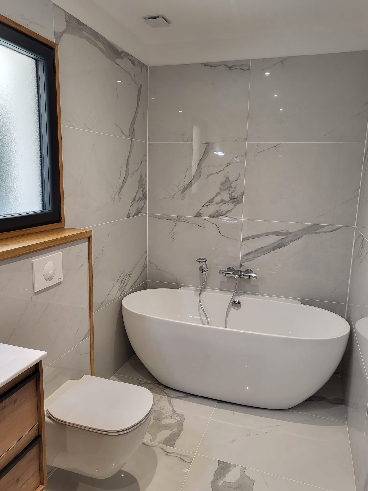 Salle de bains moderne avec baignoire îlot, carrelage en marbre gris, fenêtre, toilettes et lavabo.