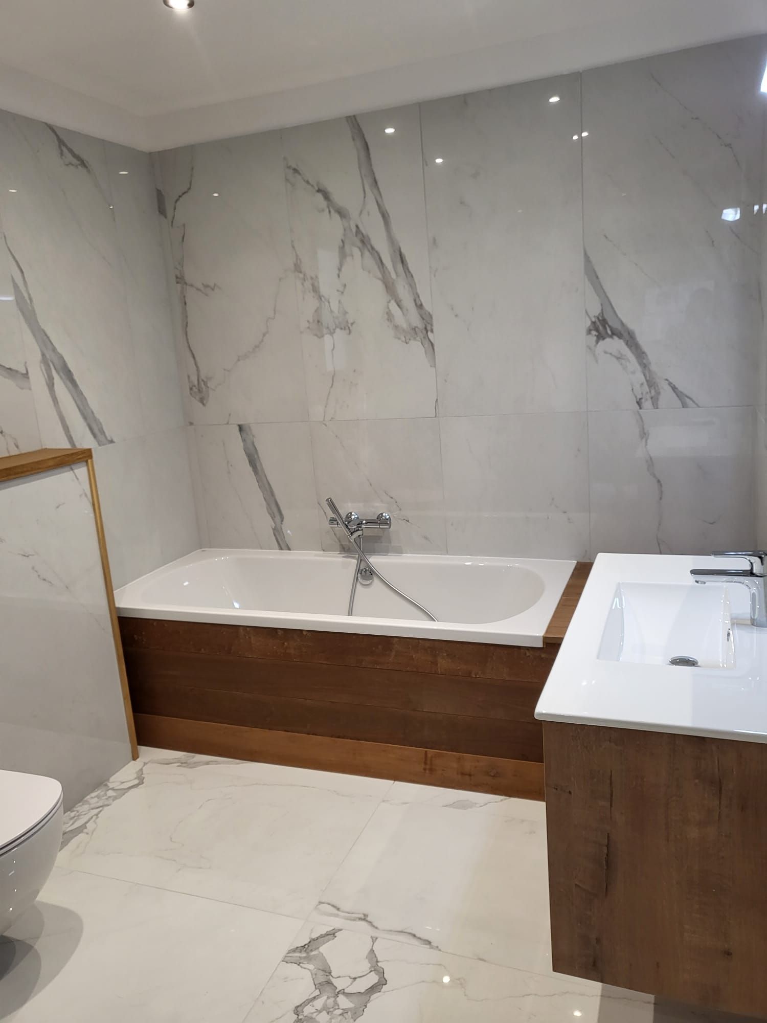 Salle de bains moderne avec murs en marbre, meuble lavabo en bois, baignoire et sol carrelé blanc.