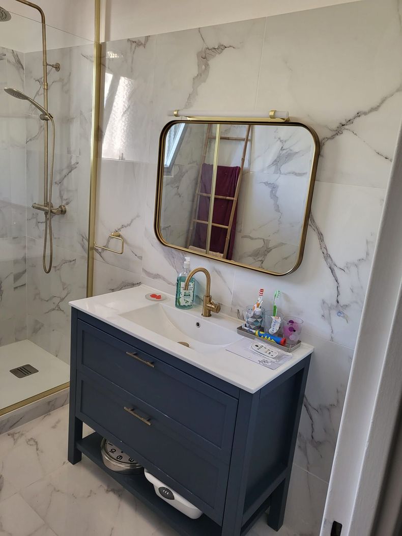 Petite salle de bains avec carrelage imitation marbre, meuble lavabo bleu marine, miroir à cadre doré et cabine de douche sur la gauche.