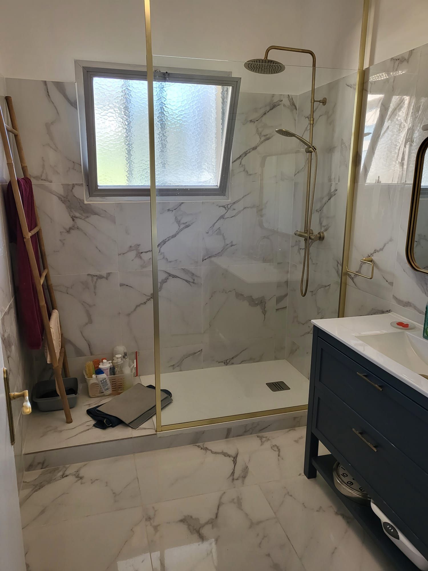 Salle de bains carrelée de marbre avec paroi de douche en verre, garnitures en laiton et tiroir de meuble lavabo foncé visible.