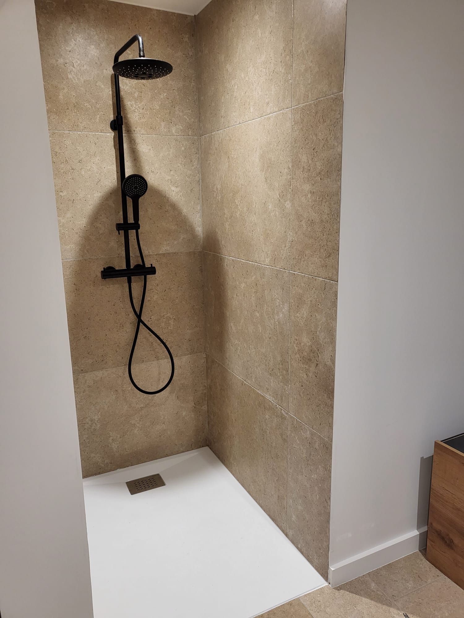 Cabine de douche d'angle avec carrelage beige, robinetterie noire et receveur de douche blanc.