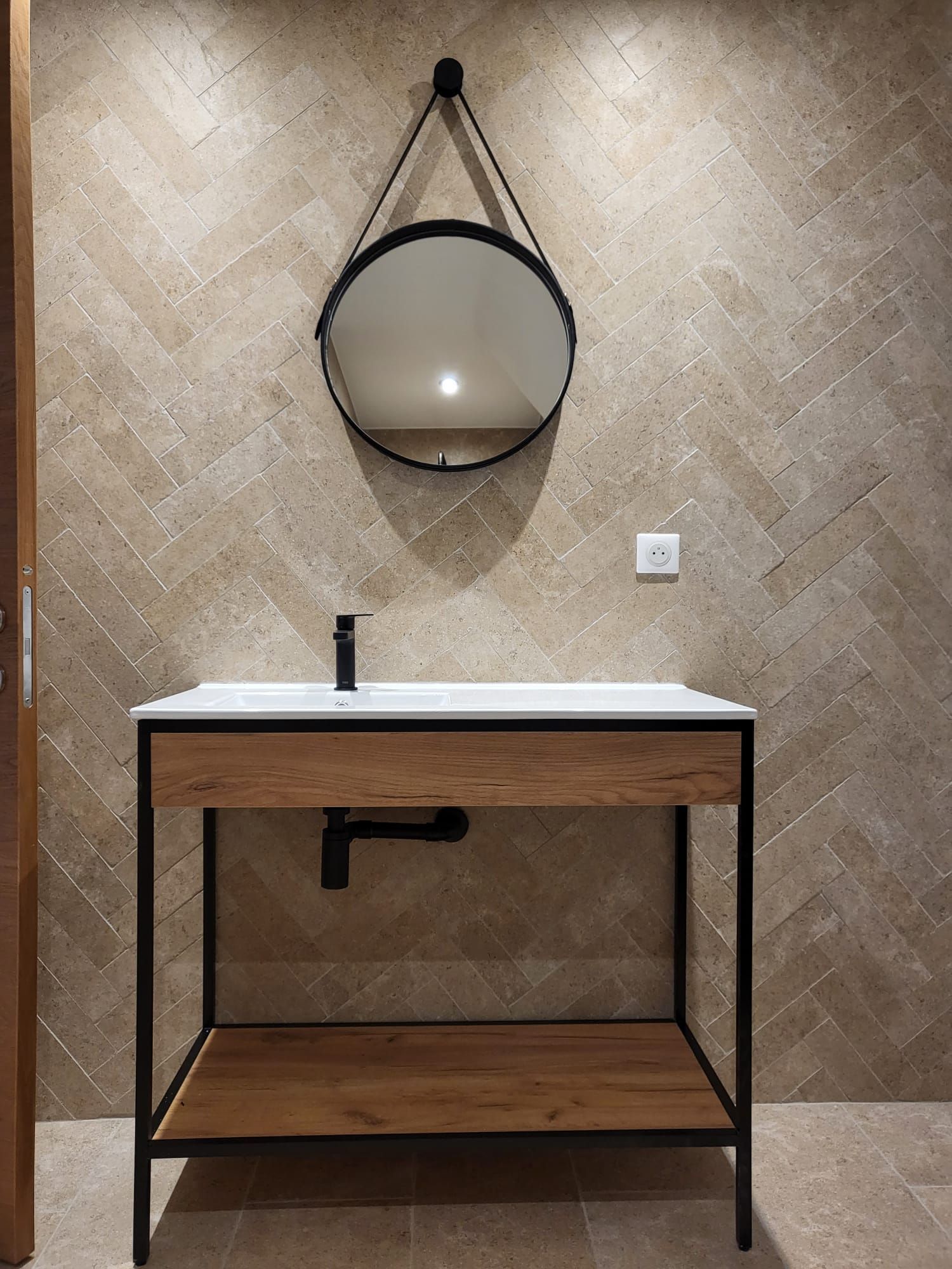 Meuble lavabo moderne avec miroir rond, mur en pierre, meuble sous-lavabo à cadre noir et étagère en bois.