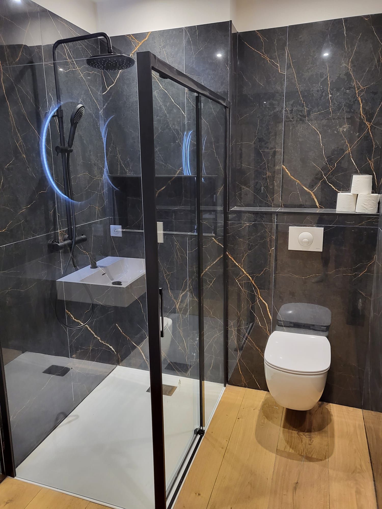 Salle de bains moderne avec douche en verre, murs en marbre noir, WC suspendu et carrelage en bois.