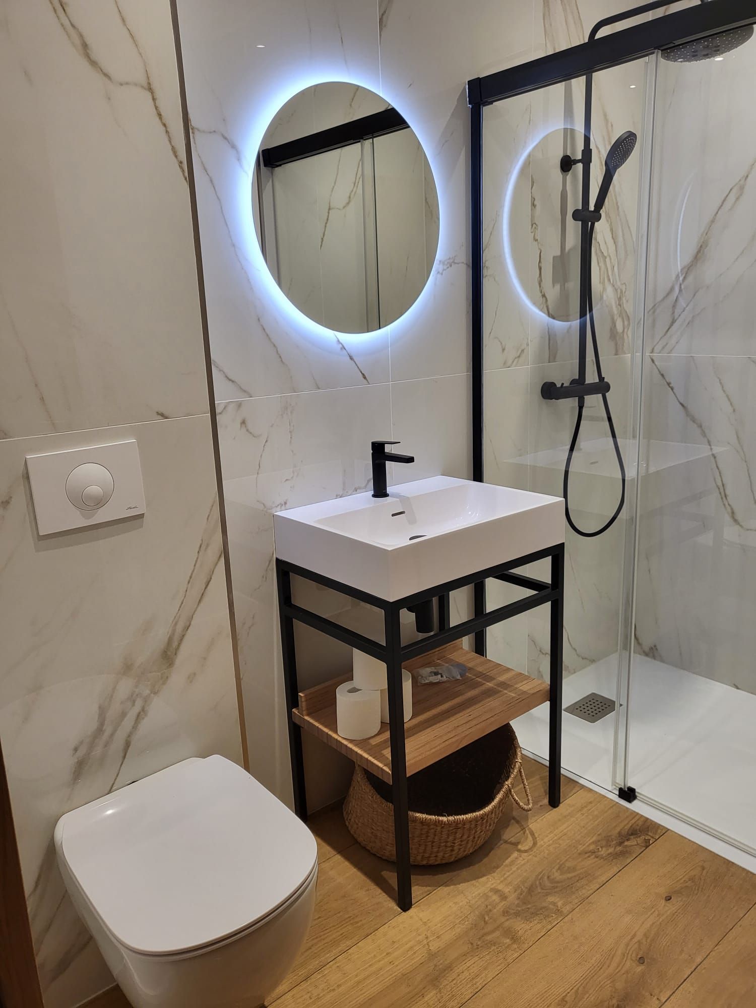 Salle de bains moderne avec toilettes suspendues, lavabo, douche et miroir rond éclairé.