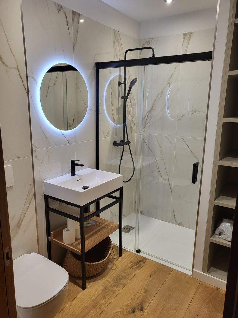 Salle de bains moderne avec miroir rond à LED, meuble lavabo noir, douche en verre et parquet.