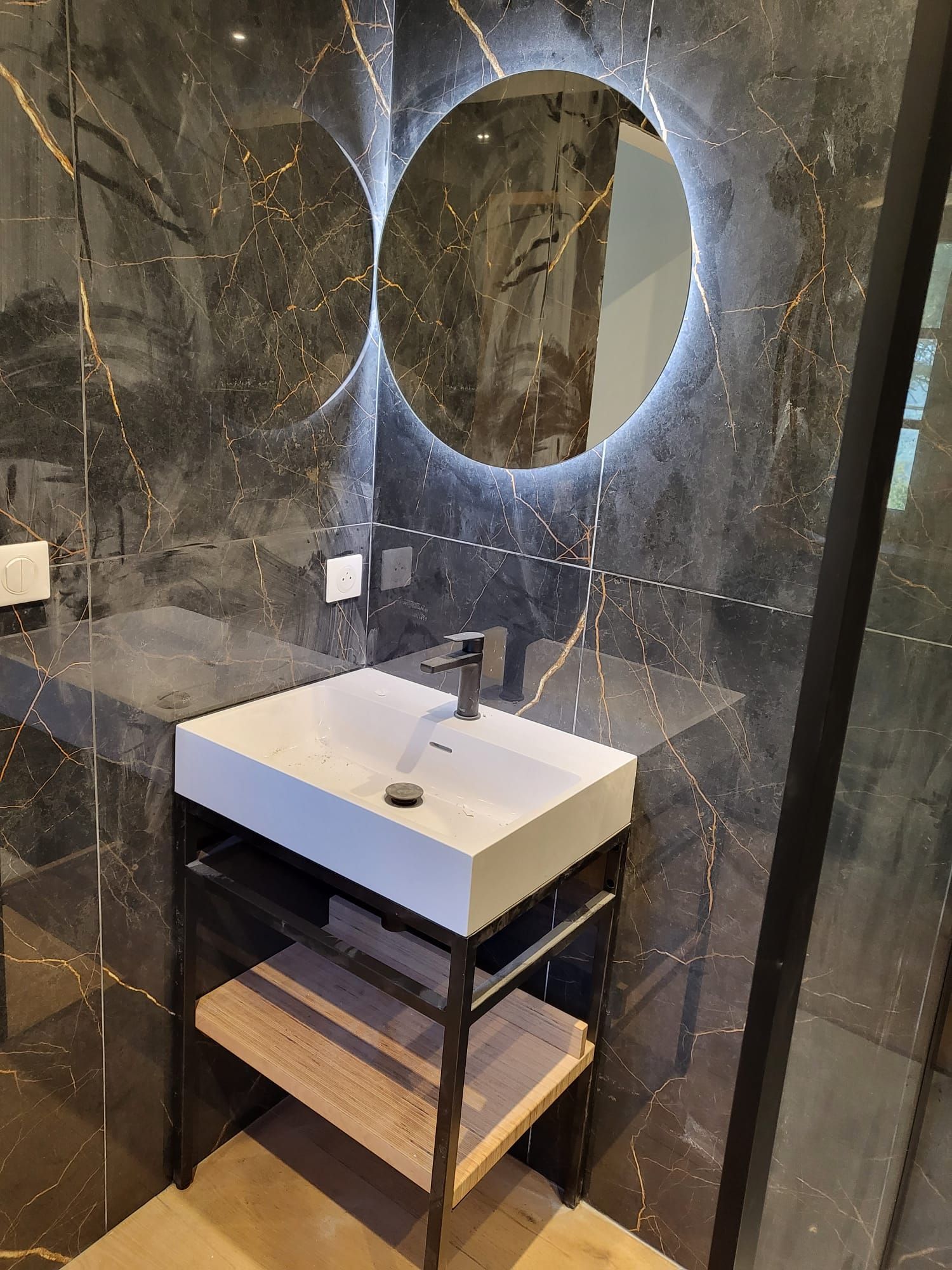 Lavabo de salle de bains moderne avec miroir rond rétroéclairé et murs carrelés en marbre foncé.