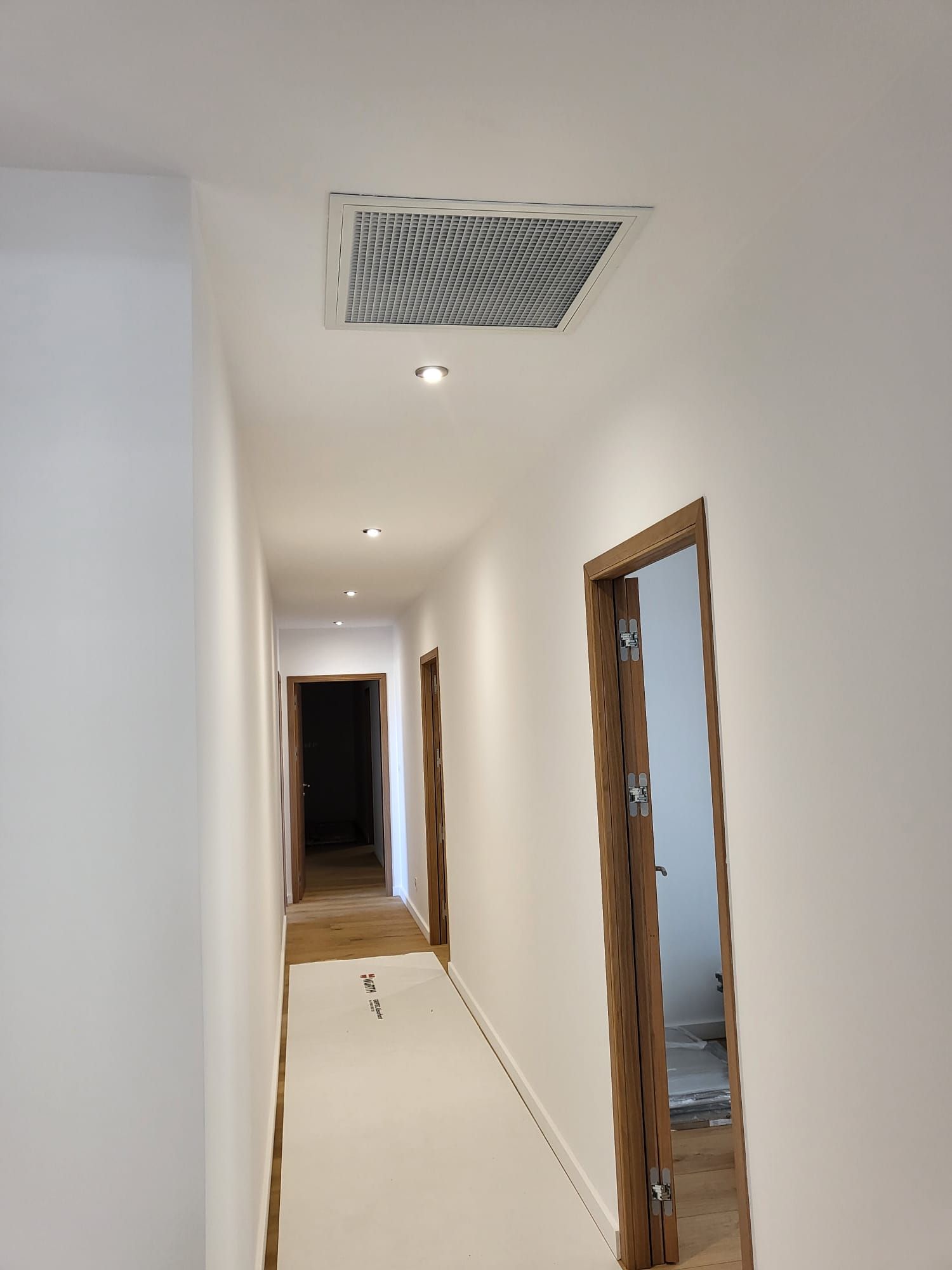 Couloir blanc étroit avec spots encastrés, grille d'aération au plafond et encadrements de portes en bois.