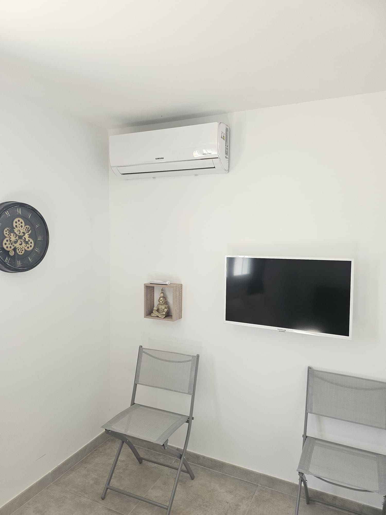Petite chambre blanche avec climatisation murale, télévision, deux chaises pliantes et un élément décoratif mural.